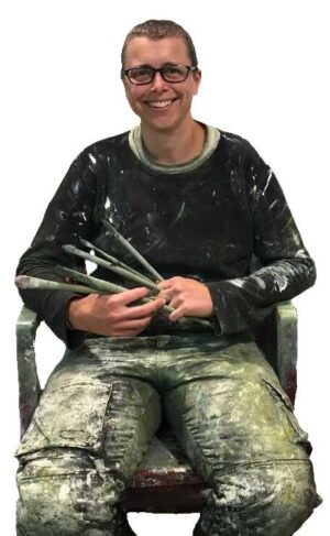 Profile photo of Urban oil painter, Leanne M Chrisie in her studio. Leanne is seated and is holding a set of oil painting brushes. Leanne is wearing dark painting clothes that are splattered by paint.